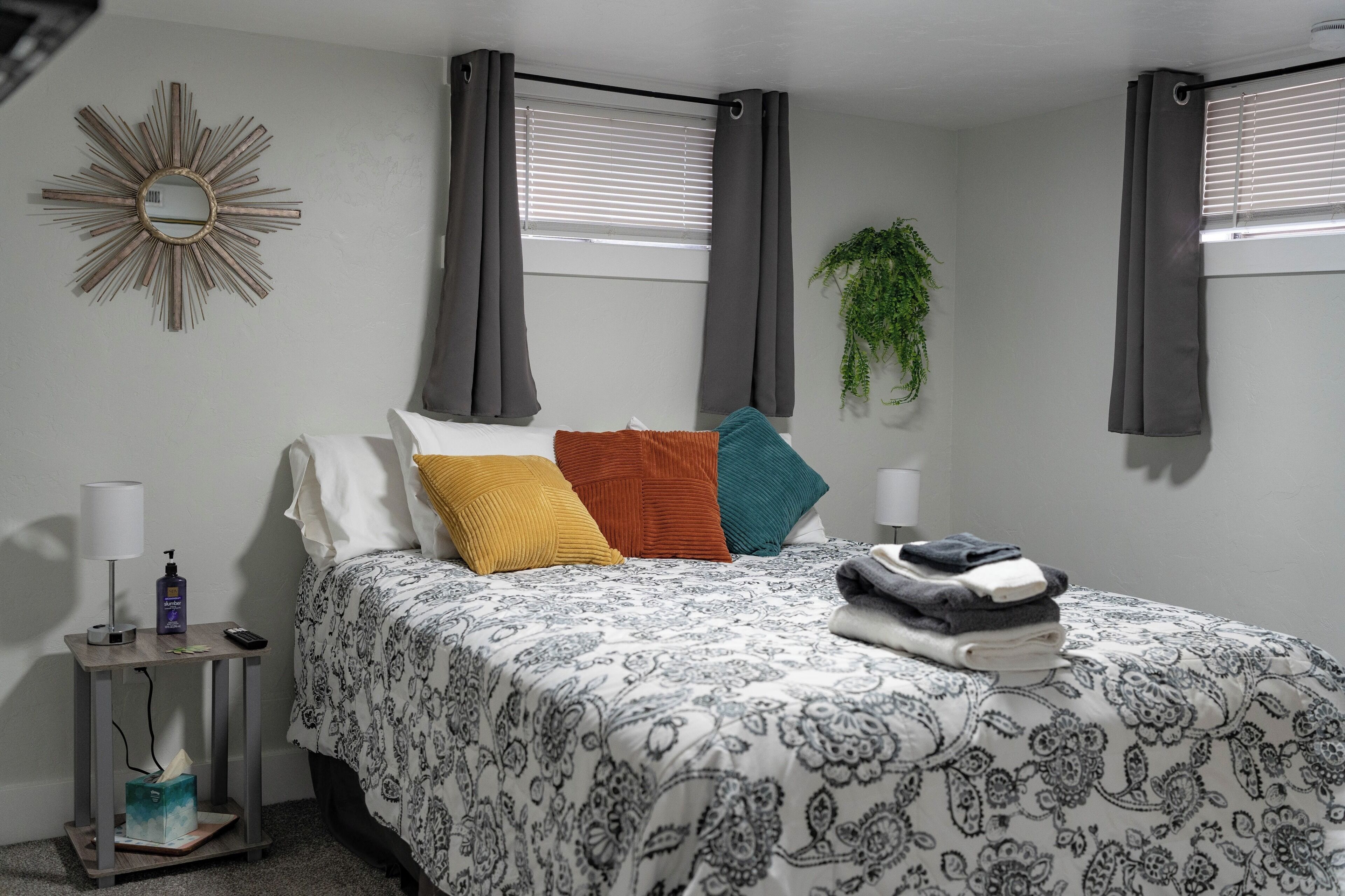 A bedroom with a bed, pillows, a nightstand, a lamp, a mirror, and a window with curtains.