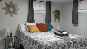A bedroom with a bed, pillows, a nightstand, a lamp, a mirror, and a window with curtains.