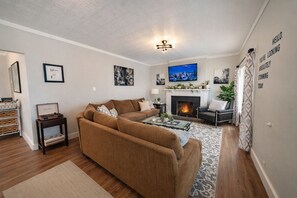 A living room with a sectional sofa, a fireplace, a flat-screen TV, and a wall-mounted artwork. - Guest Favorite! Hot Tub, Huge Kitchen, Fenced Yard, Firepit, & BBQ in Great Area (Price)