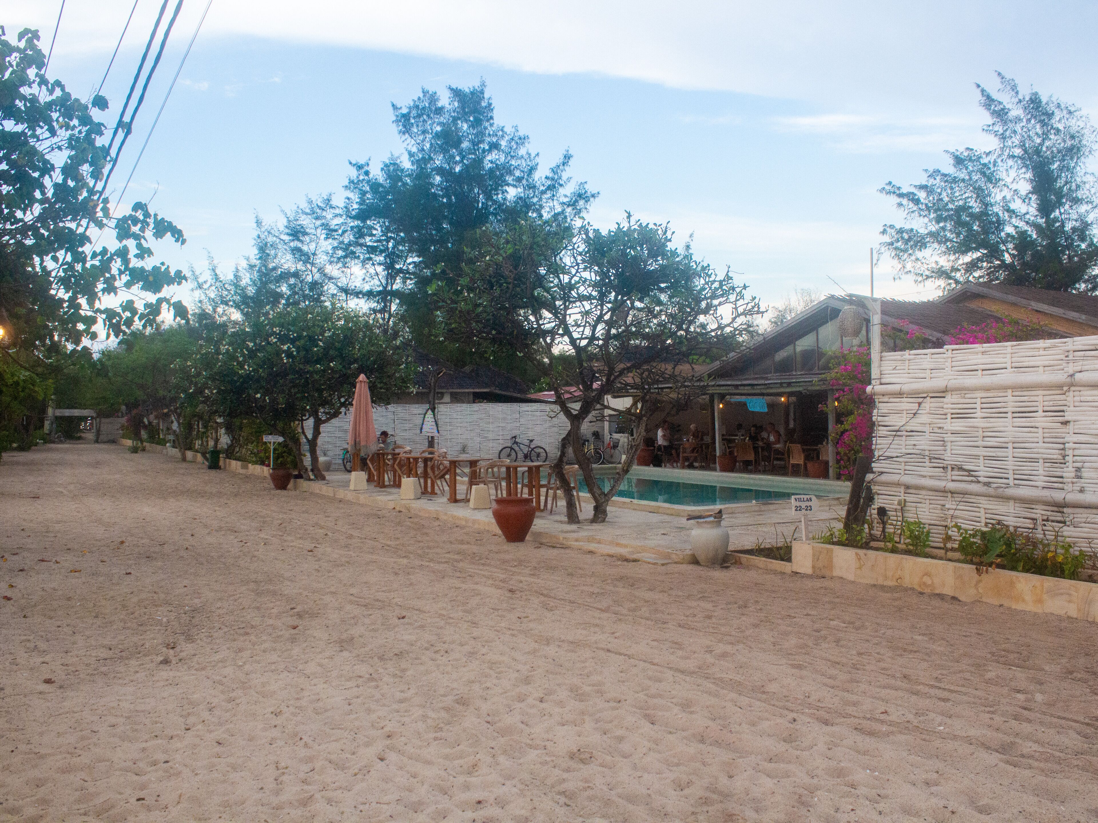 A sandy outdoor area with a pool, wooden furniture, and a covered seating area.