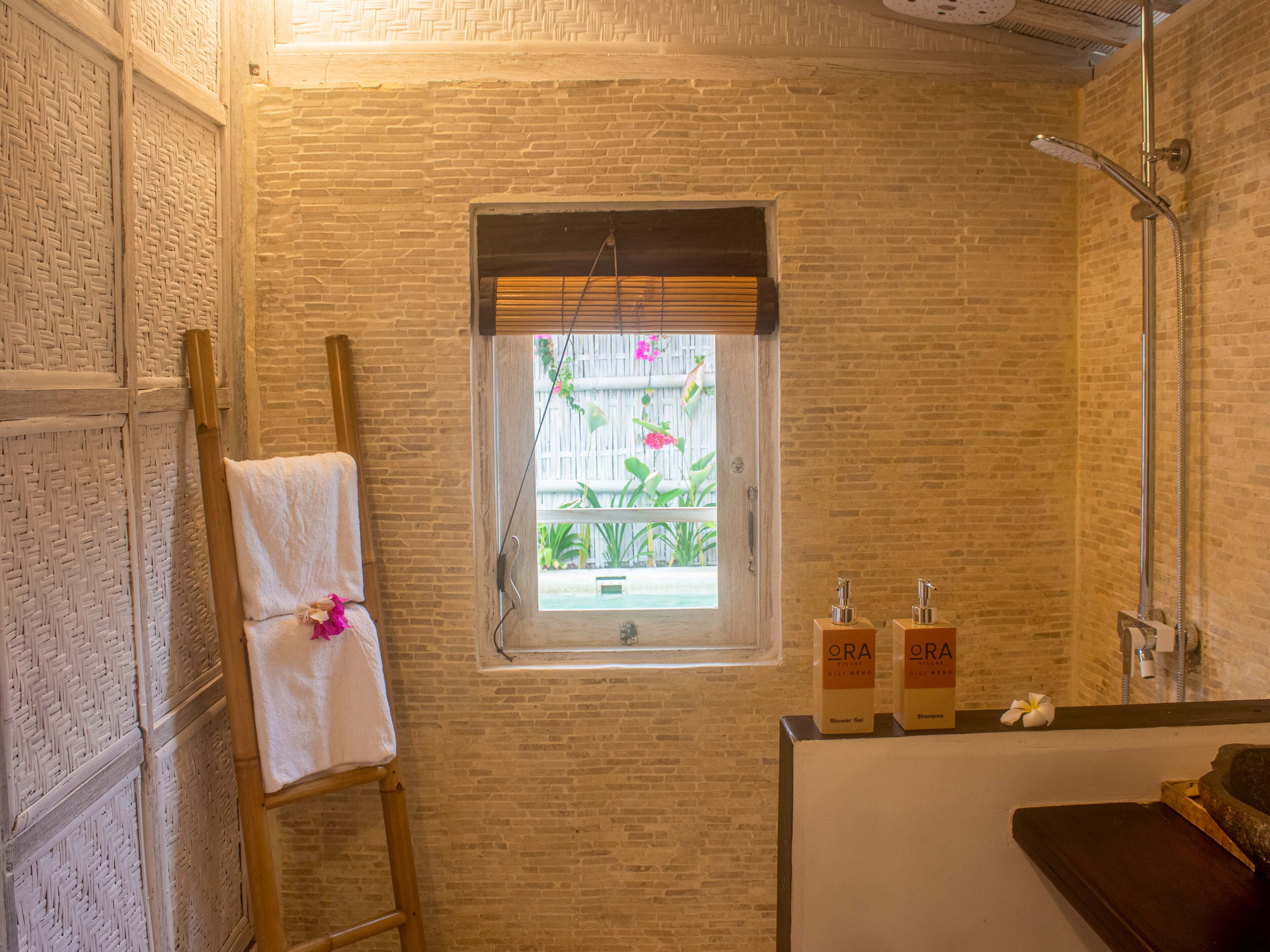 A bathroom with a brick wall, a window with a view of plants, a towel on a wooden stool, and toiletries on a counter.