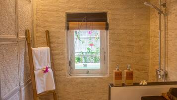 A bathroom with a brick wall, a window with a view of plants, a towel on a wooden stool, and toiletries on a counter.