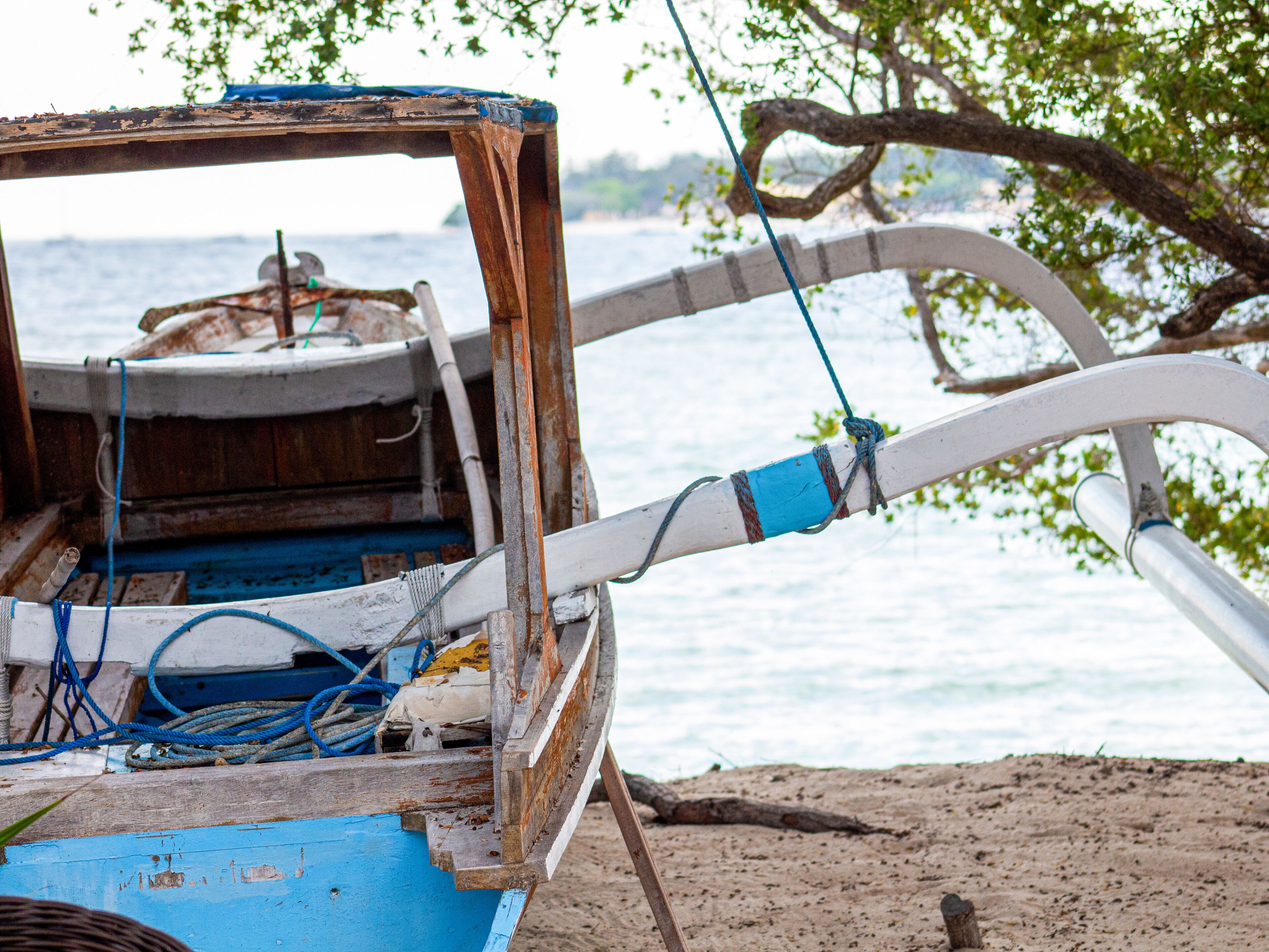 Un bateau dont le pont est usé et dont les cordes sont bien visibles, ancré près d’une plage de sable, avec une étendue d’eau en arrière-plan.