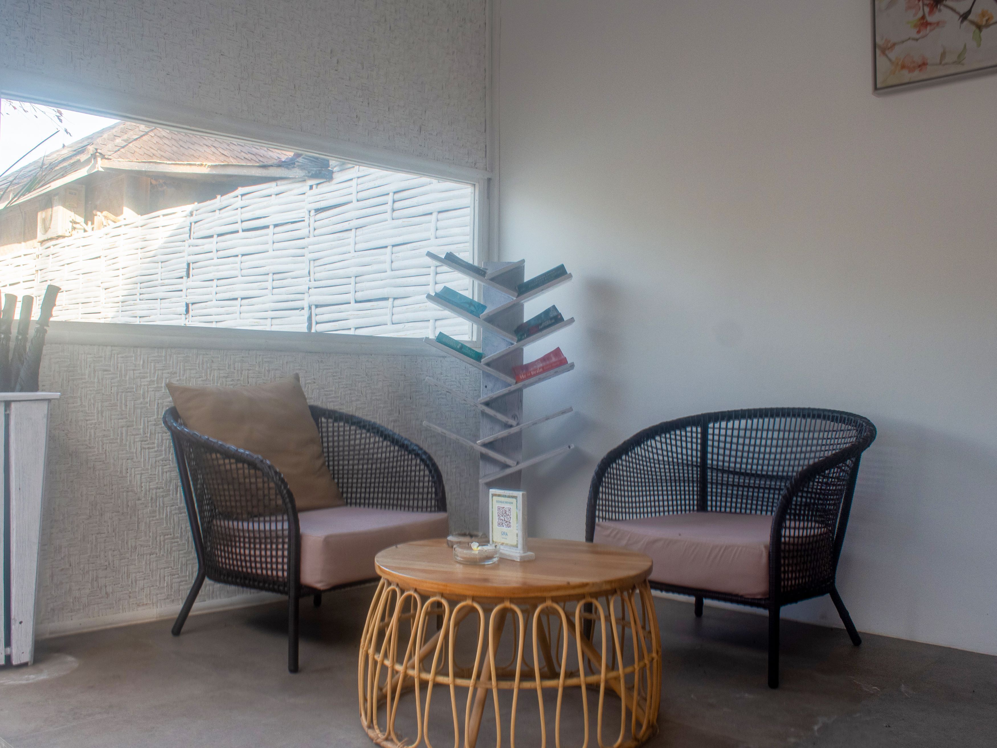 Un coin salon accueillant avec deux fauteuils en osier et une table ronde, une bibliothèque remplie de livres et une fenêtre donnant sur l’extérieur.
