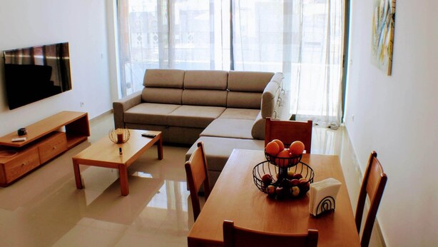 A living room with a sofa, a TV, a wooden coffee table, and a dining table with fruit and a napkin. - Kingfisher Plateau. 2A Apt on Rua Pedonal. Praia Central_ Near cafes & shops (Praia)