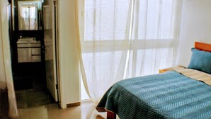 A bedroom with a bed, a nightstand, a window with sheer curtains, and a door leading to a bathroom. - Kingfisher Plateau. 2A Apt on Rua Pedonal. Praia Central_ Near cafes & shops (Praia)