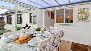 A conservatory with a dining table set for four, surrounded by white chairs. There is a large window on one side and a wall with a decorative sign. - Kittiwake - Short Walk to Beach and Village (Saundersfoot)