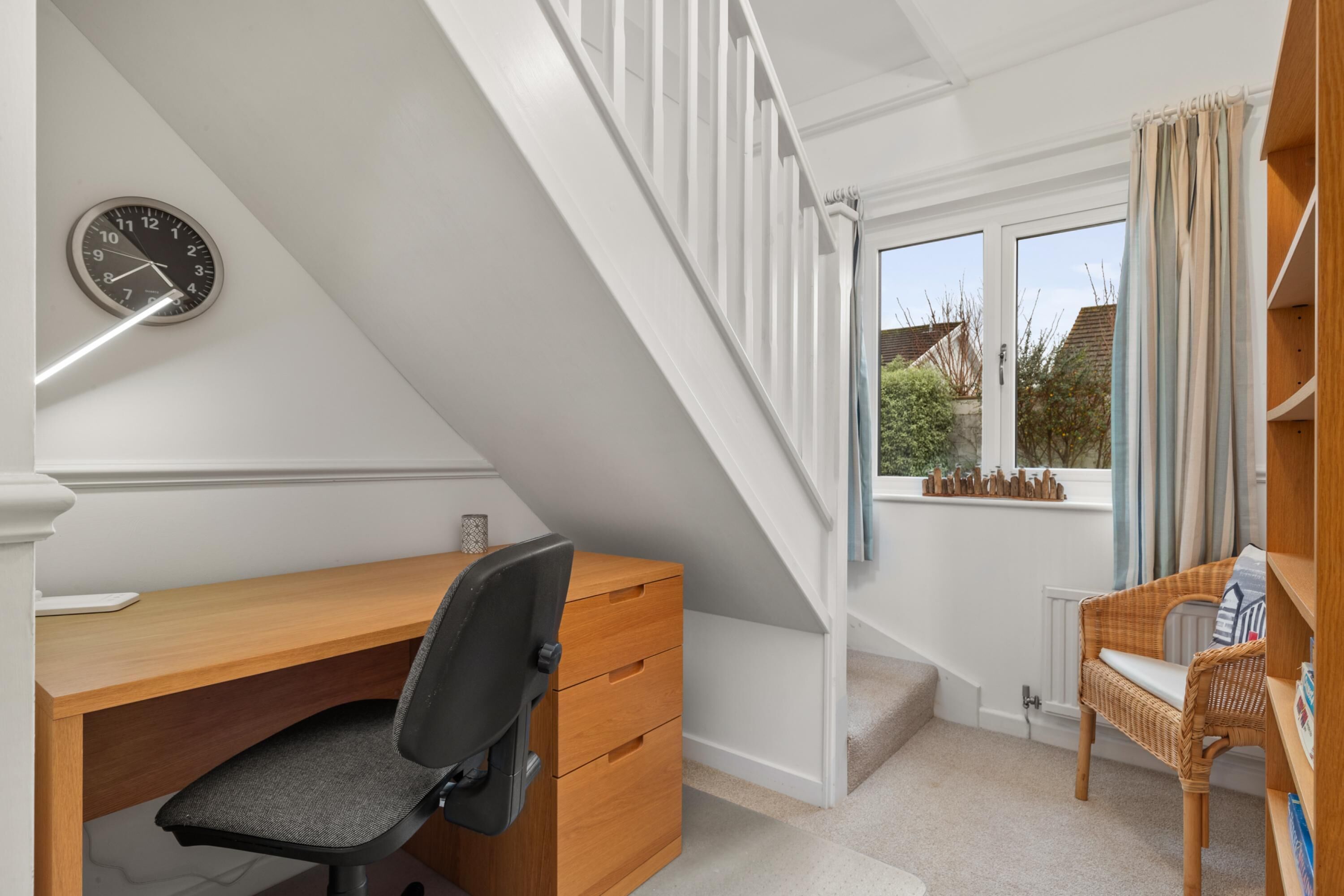 Une pièce avec un bureau en bois, un fauteuil de bureau noir, une horloge murale, une fenêtre avec des rideaux et un escalier.