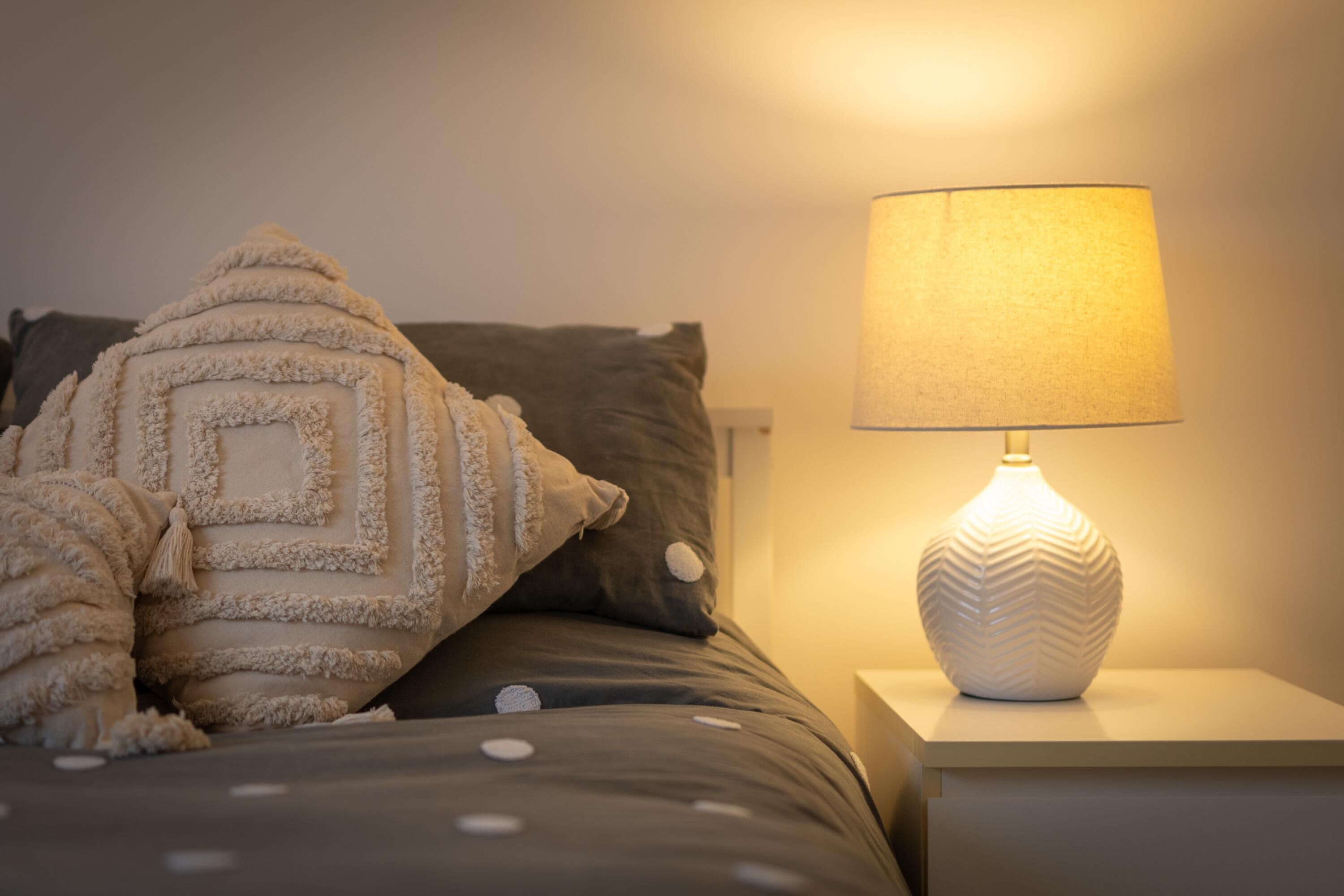 Una cama con una manta y almohadas estampadas, una mesita de noche blanca con una lámpara y una pared con interruptor de luz.