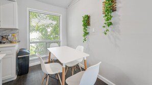 Une cuisine moderne avec des meubles blancs, une table blanche et des chaises. On y trouve une fenêtre avec des stores, une poubelle et une plante accrochée au mur.