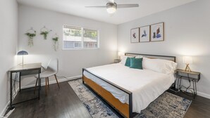 Une chambre à coucher comprenant un lit, un bureau, une chaise, un ventilateur de plafond, une fenêtre avec stores, des plantes fixées au mur et des œuvres d’art encadrées.