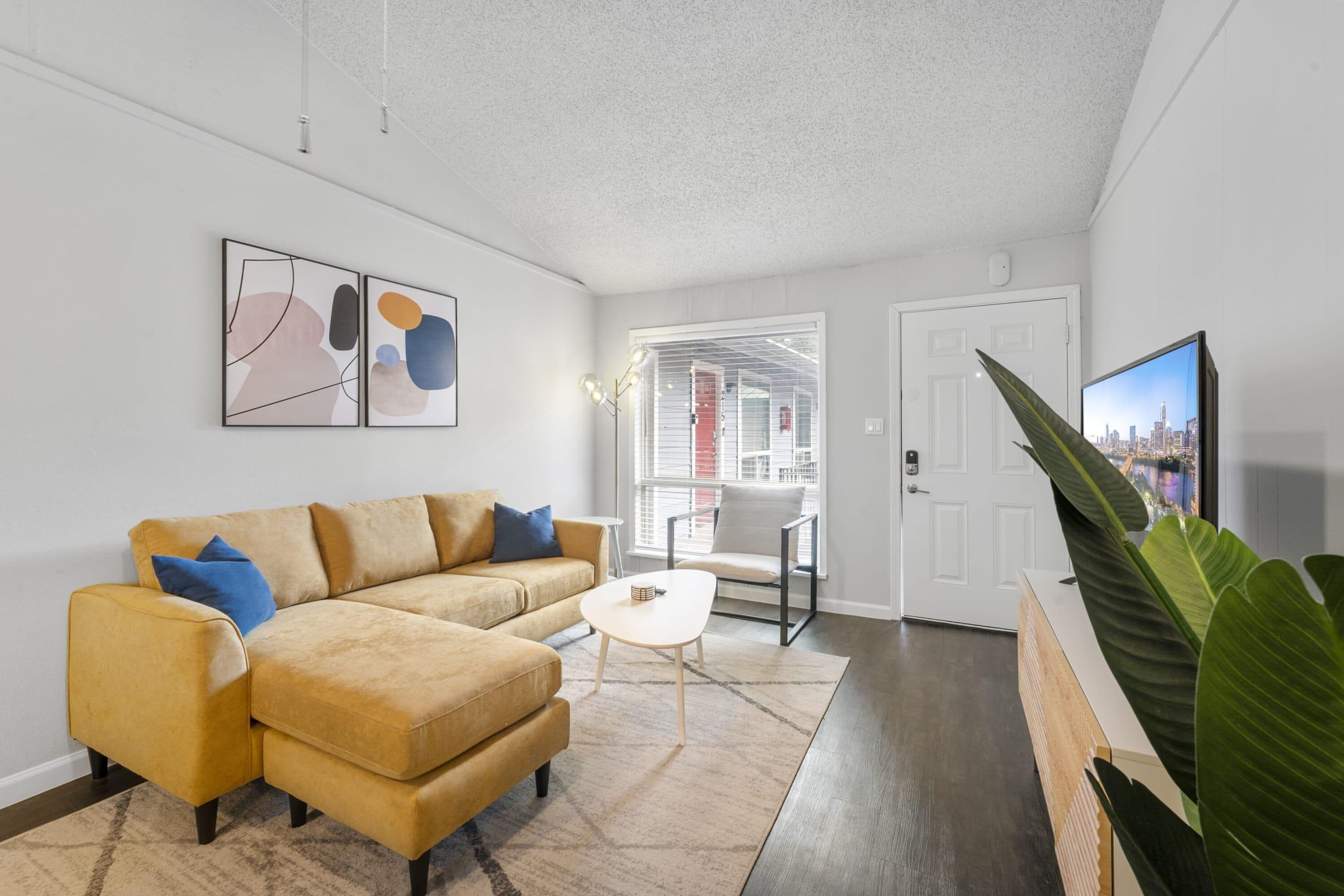 Een moderne woonkamer met een beige modulaire bank, een kleine witte salontafel en een grote plant. Er is een deur die leidt naar een balkon met uitzicht op de stad.