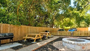 Un jardin avec une clôture en bois, un barbecue, des tables de pique-nique et un foyer en pierre.
