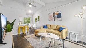 A modern living room with a sofa, coffee table, and TV. There is a dining area with yellow stools and a white table. The room has white walls and a ceiling fan. - Prime Soco Comfy 2BR King Suite w Pool Parking (Austin)