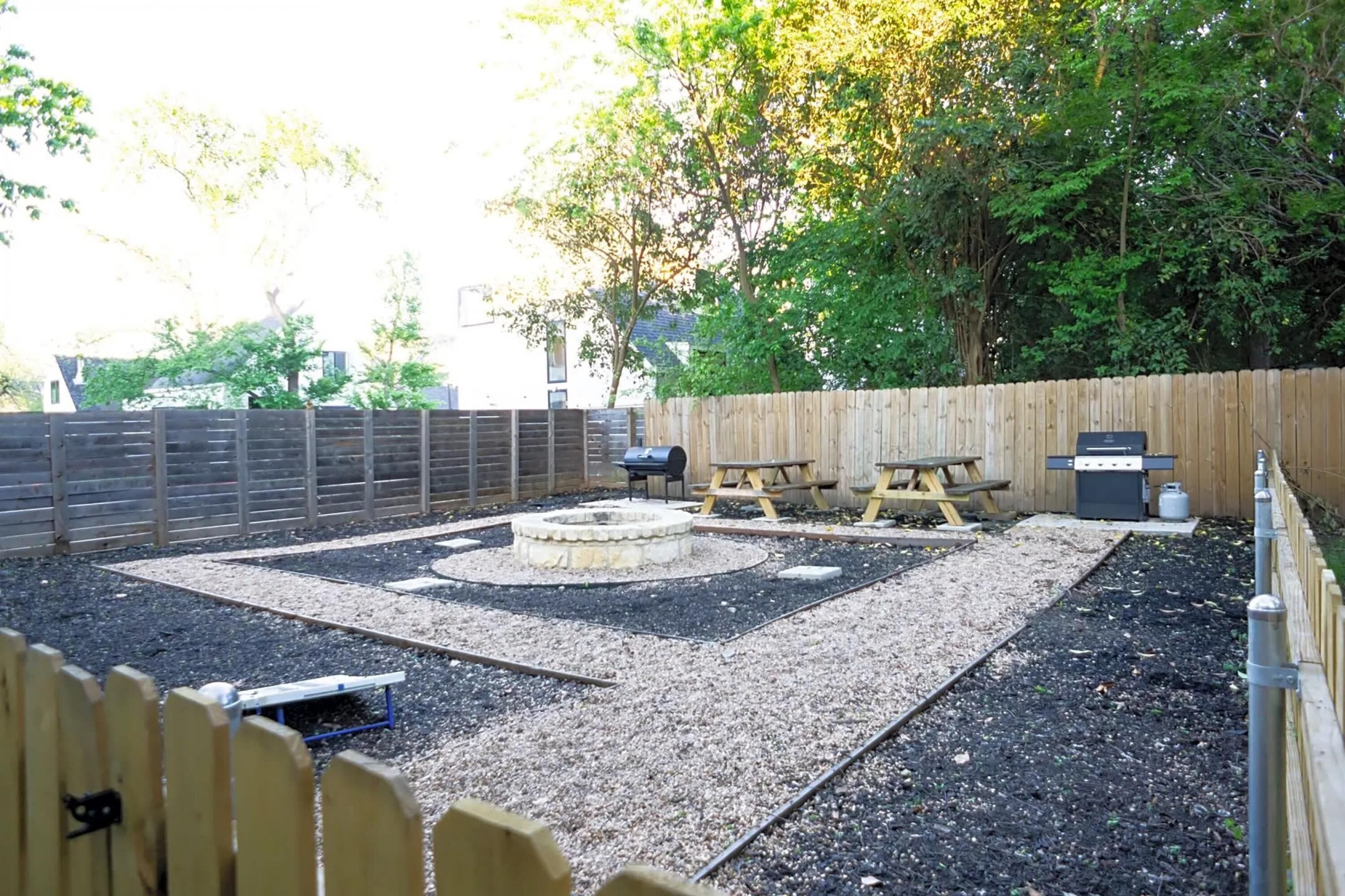 A backyard with a wooden fence, a circular fire pit, picnic tables, and a barbecue grill.