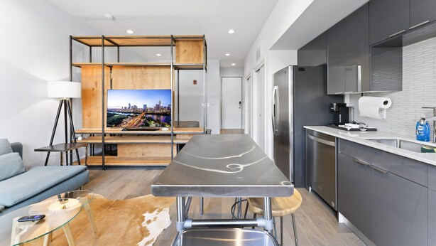 Une cuisine moderne équipée d’une cuisinière intégrée, d’un réfrigérateur et d’un évier. On y trouve une table à manger, et l’écran de télévision offre une vue sur le paysage urbain.
