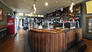 A bar area with a wooden counter, shelves with bottles, and a fan on the wall. - Royal Hotel Capertee (Capertee)