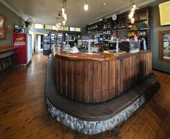 Uma área de bar com balcão de madeira, prateleiras com garrafas e um ventilador na parede.