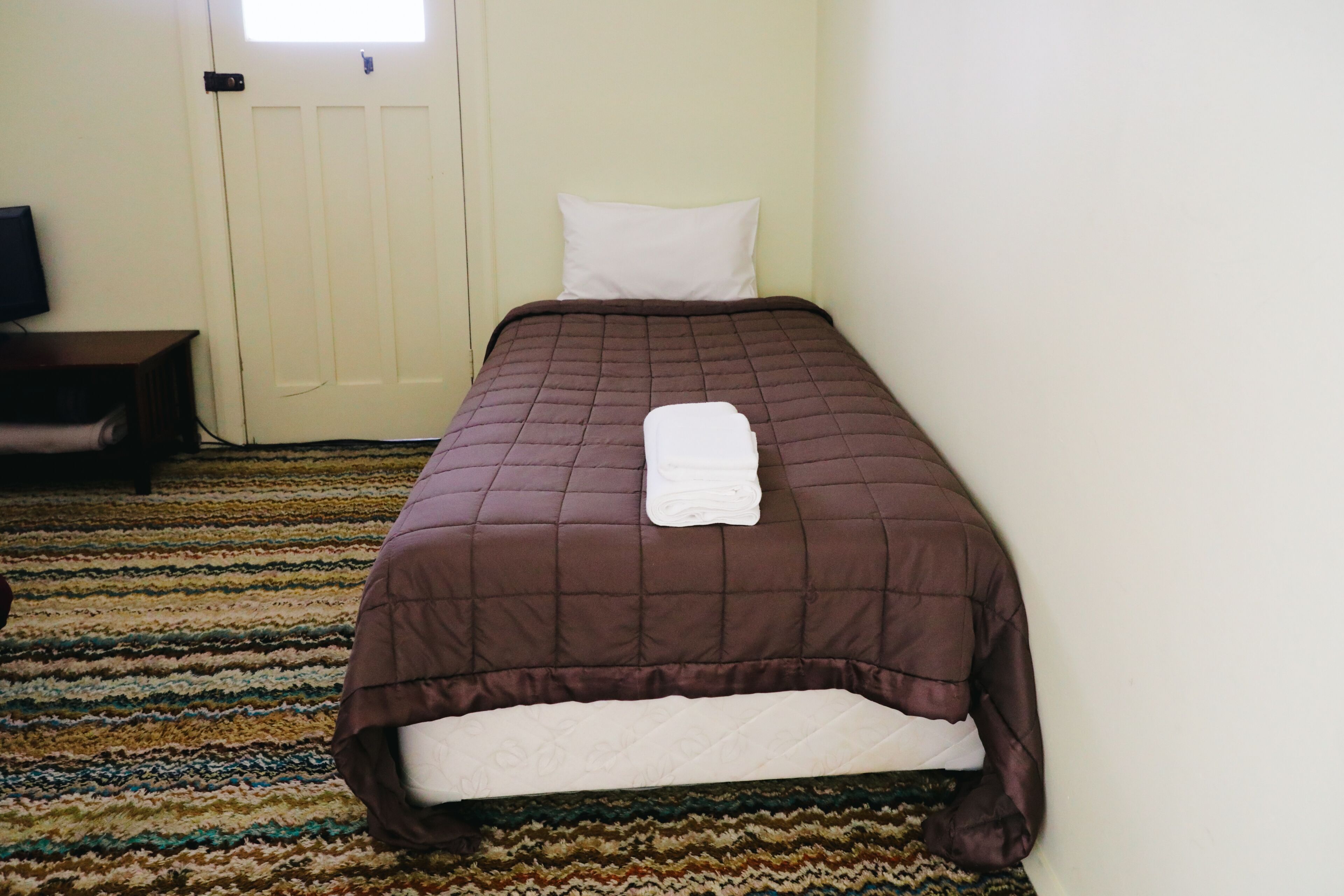 A neatly made bed with a brown quilt and white pillows, a television on a wooden stand, and a patterned carpet.
