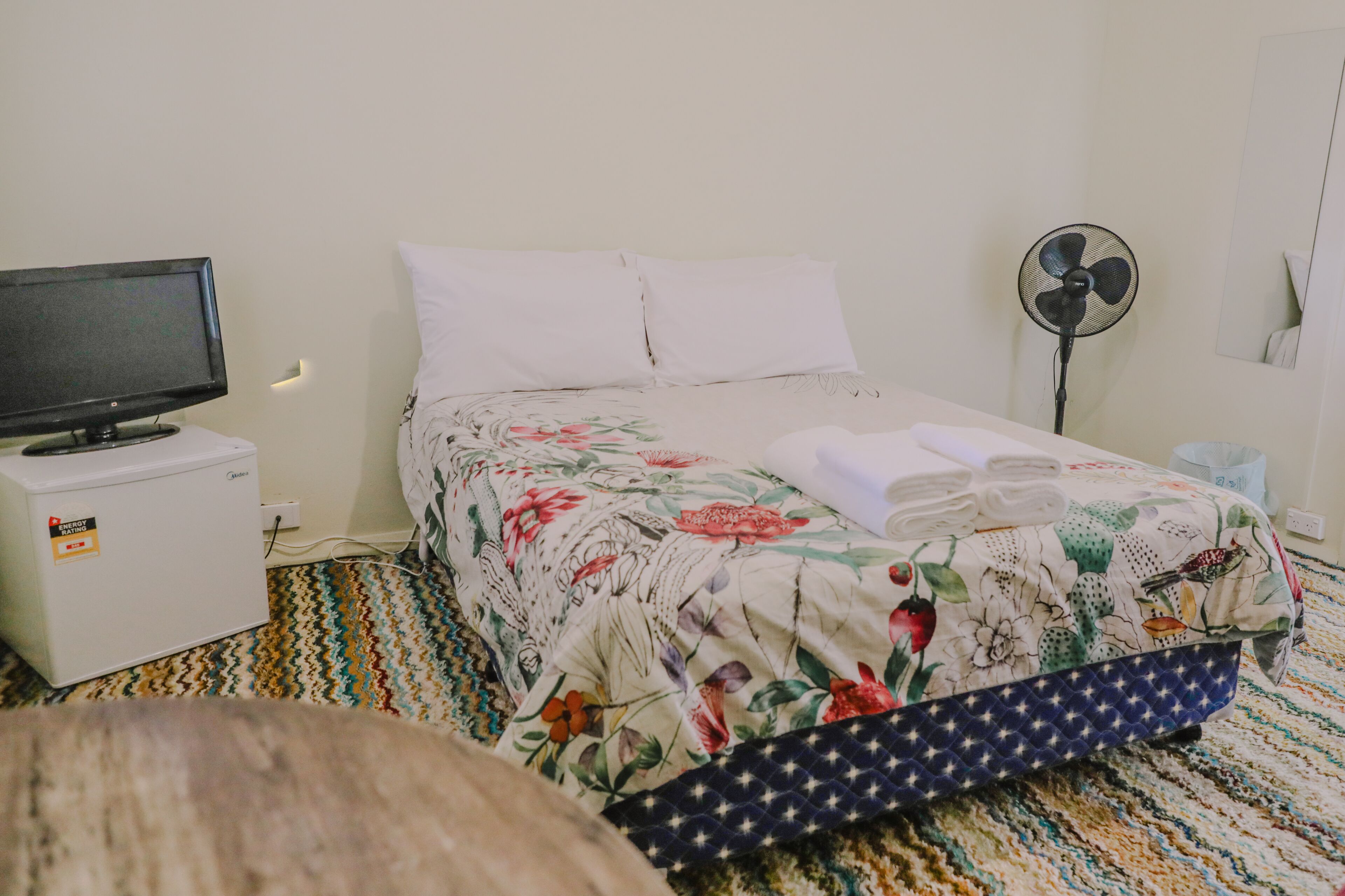 A hotel room with a bed, a television on a nightstand, a fan, and a small table with a power strip.