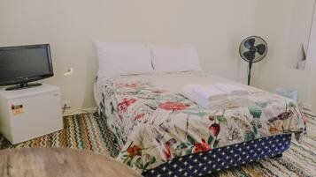 A hotel room with a bed, a television on a nightstand, a fan, and a small table with a power strip.