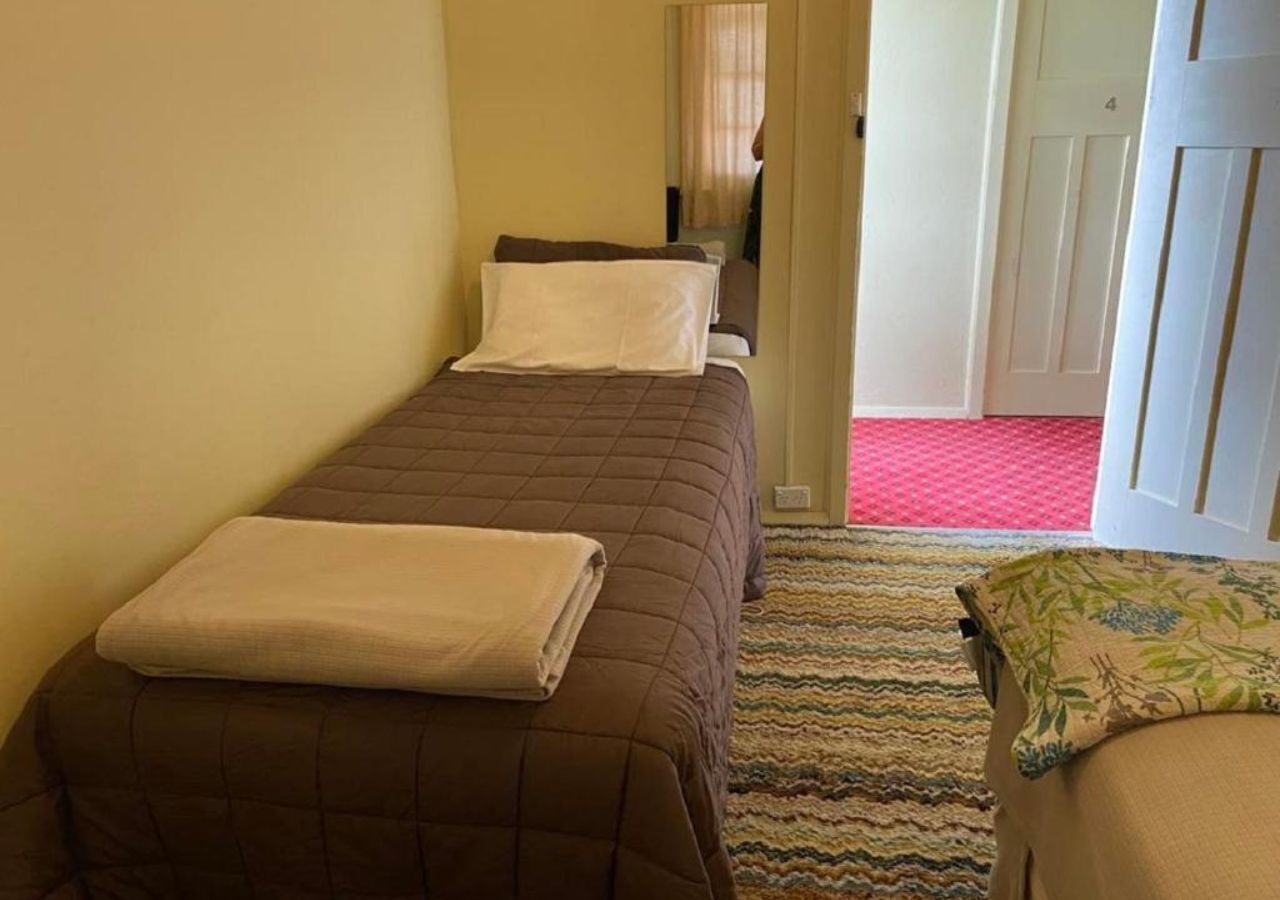 Una habitación pequeña y estrecha con dos camas individuales, cada una con una colcha marrón y almohadas blancas. Una alfombra a rayas cubre el suelo, y se ve una puerta al fondo.