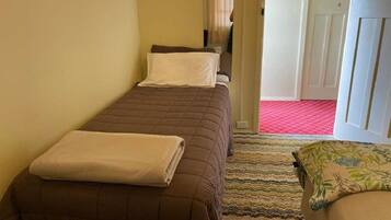 Una habitación pequeña y estrecha con dos camas individuales, cada una con una colcha marrón y almohadas blancas. Una alfombra a rayas cubre el suelo, y se ve una puerta al fondo.
