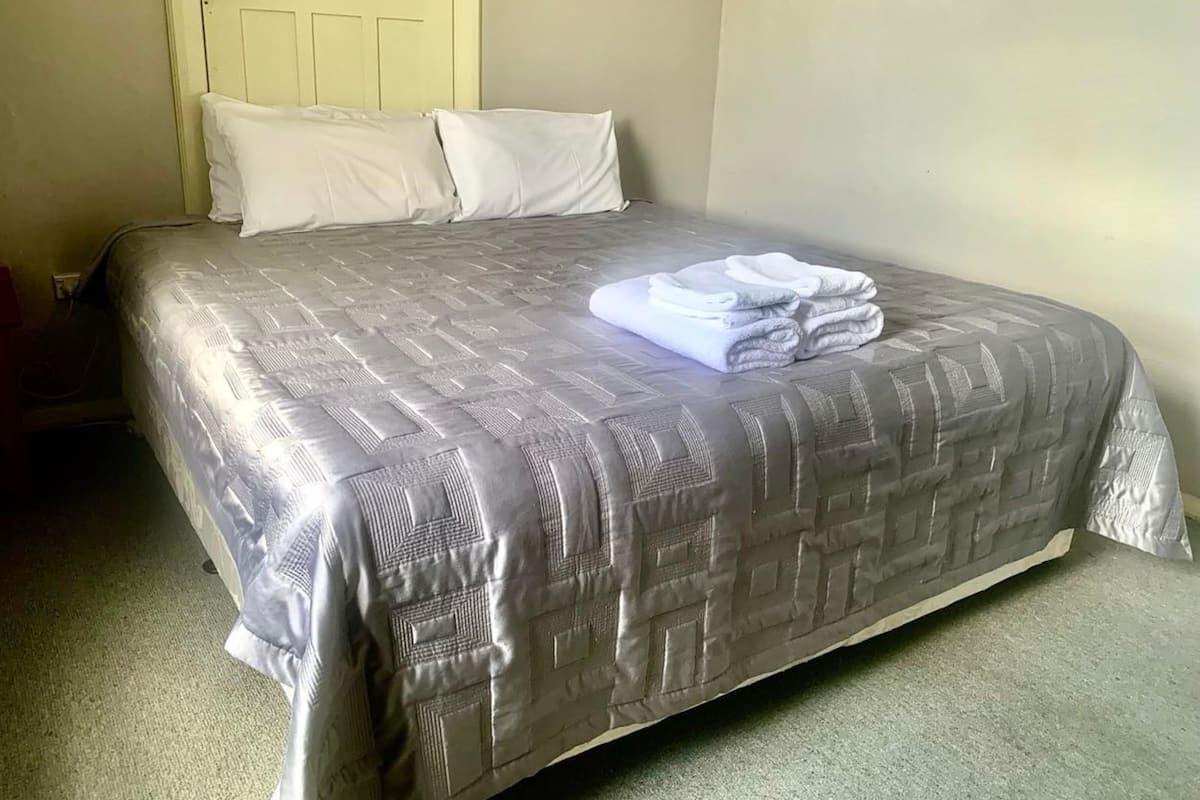 Ein ordentlich bezogenes Bett mit einem gemusterten Überwurf und Kissen, ein gefaltetes Handtuch auf dem Bett sowie eine Tür mit Griff und Lichtschalter.