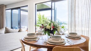 A dining area with a round table set for four, a sofa, and large windows with blinds. - Super Central in Wanaka - Luxury villa only a 10 minute walk to town (Wanaka)
