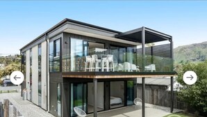 A modern two-story house with a balcony, glass doors, and a small patio area with chairs. - Super Central in Wanaka - Luxury villa only a 10 minute walk to town (Wanaka)