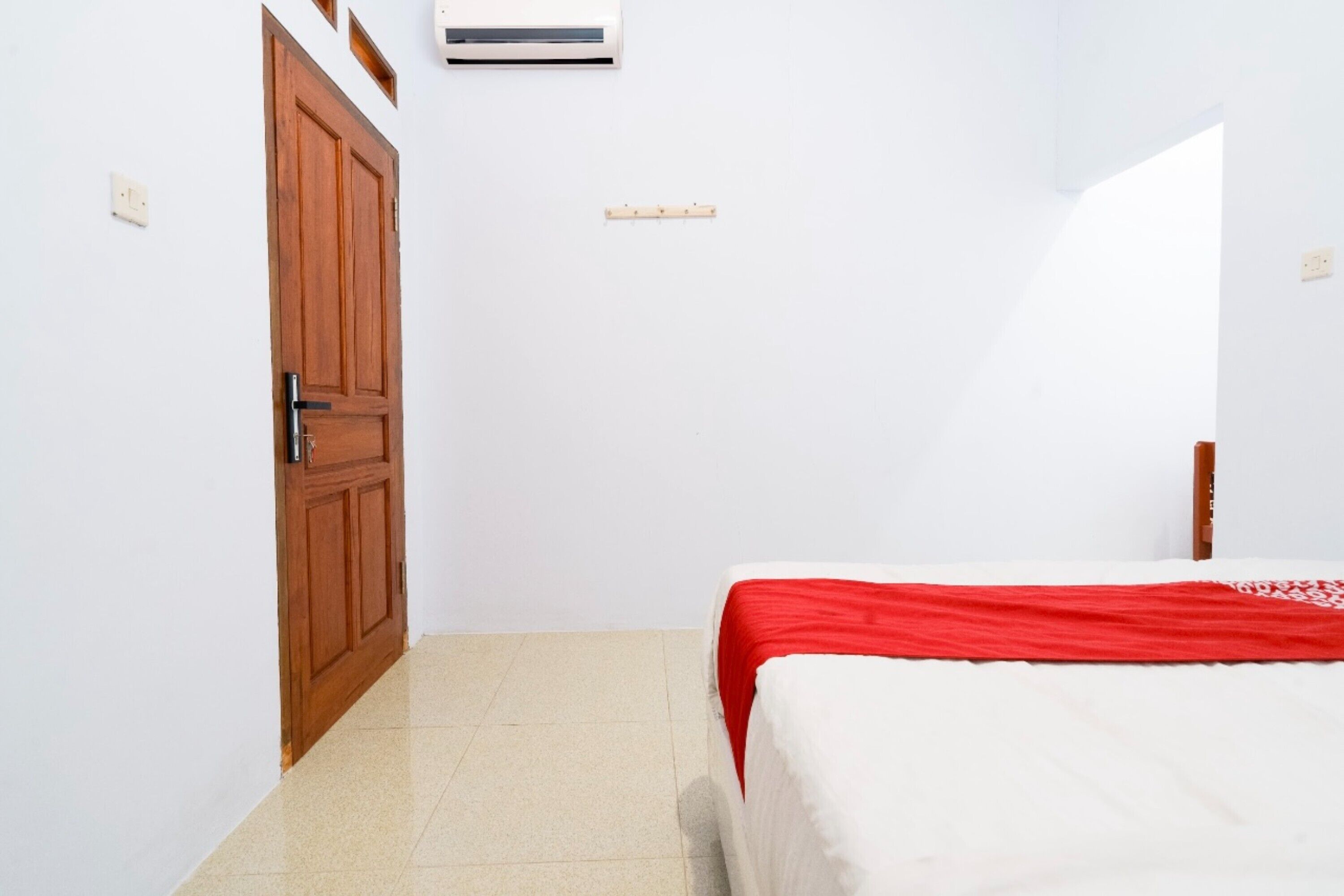 A small, clean room with a wooden door, a bed with a red and white bedspread, and a white air conditioning unit on the wall.