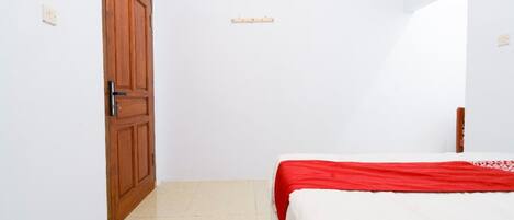 A small, clean room with a wooden door, a bed with a red and white bedspread, and a white air conditioning unit on the wall.
