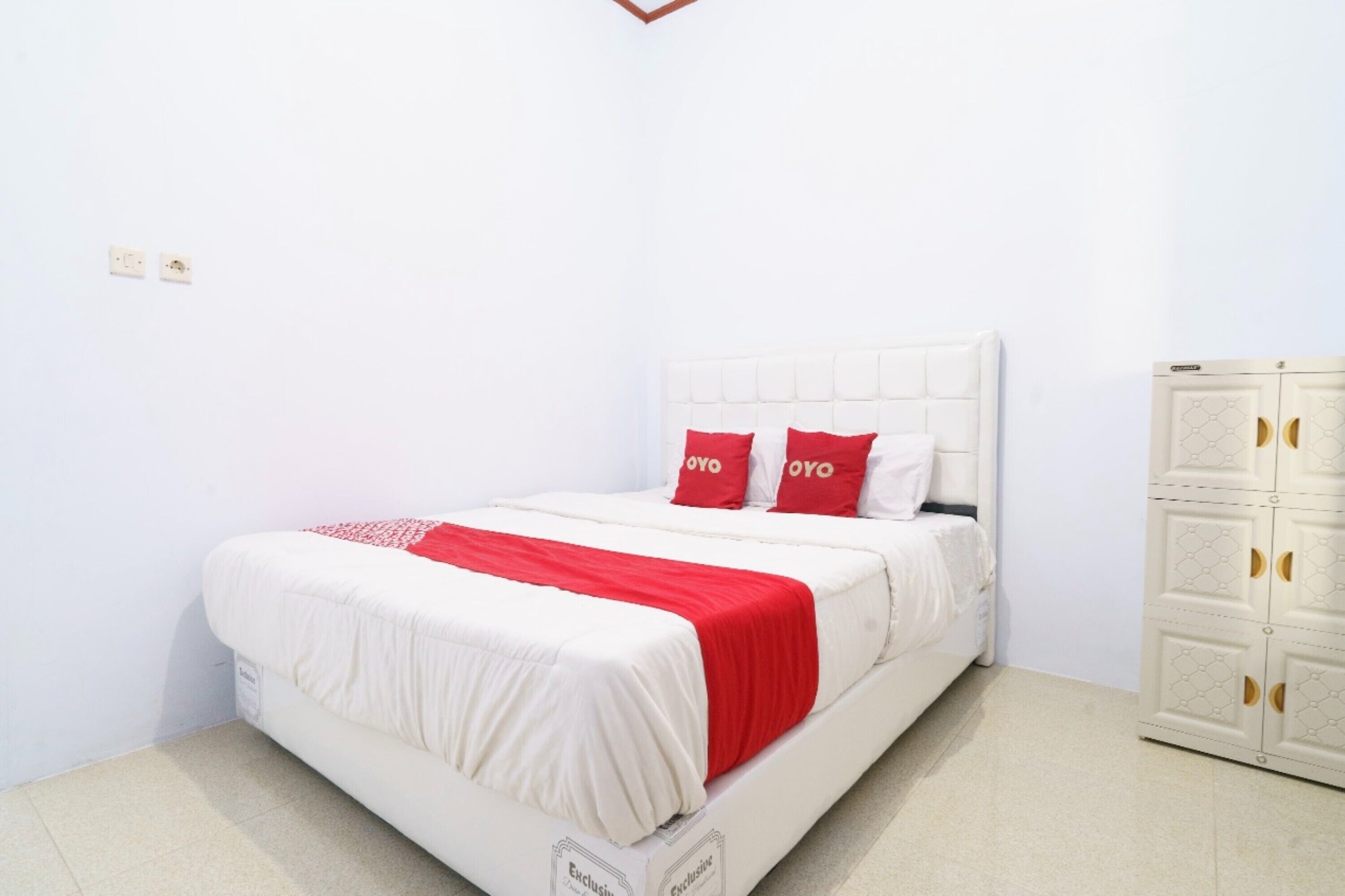 A neatly made bed with a red and white bedspread and two red pillows, a white headboard, and a white wardrobe with a geometric pattern.