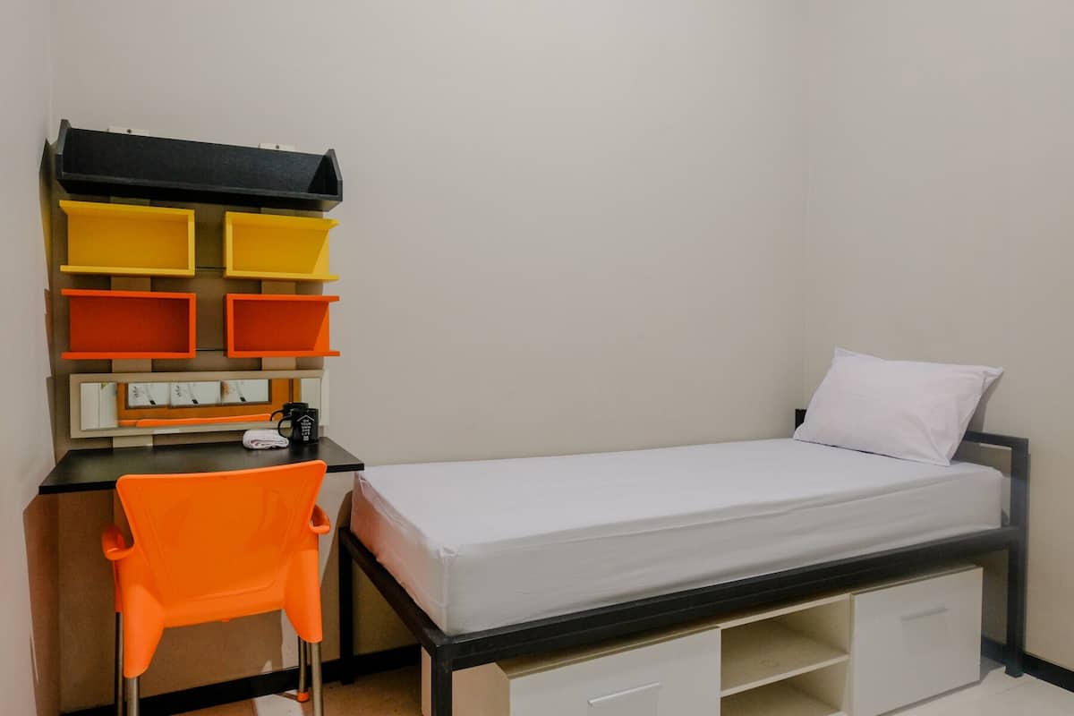 Ein kleines Zimmer mit einem Einzelbett, einem Schreibtisch, einem Stuhl, einem Bücherregal und einem weißen Kopfkissen.
