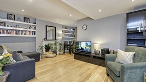 A modern living room with a flat-screen TV, bookshelves, a clock, and a comfortable seating area. - Spacious Pimlico Apartment by Underthedoormat (London)