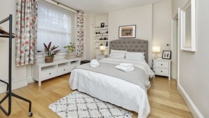 A bedroom with a bed, bedside tables, a nightstand, a lamp, a shelf with books, and a window with curtains. - Spacious Pimlico Apartment by Underthedoormat (London)