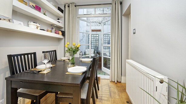 A dining area with a dark wooden table set for four, flanked by white shelves with various items. A window with curtains leads to an outdoor area. - Spacious Pimlico Apartment by Underthedoormat (London)