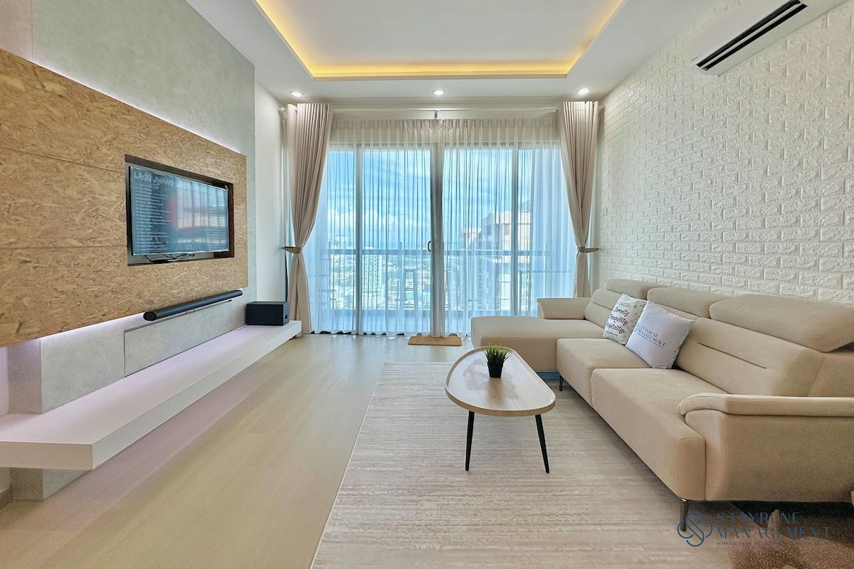 Una sala de estar moderna con un televisor de pantalla plana montado en una pared texturizada, un sofá color beige, una mesa de centro y un ventanal con cortinas translúcidas.