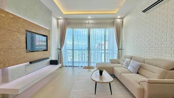 A modern living room with a flat-screen TV mounted on a textured wall, a beige sofa, a coffee table, and a large window with sheer curtains.