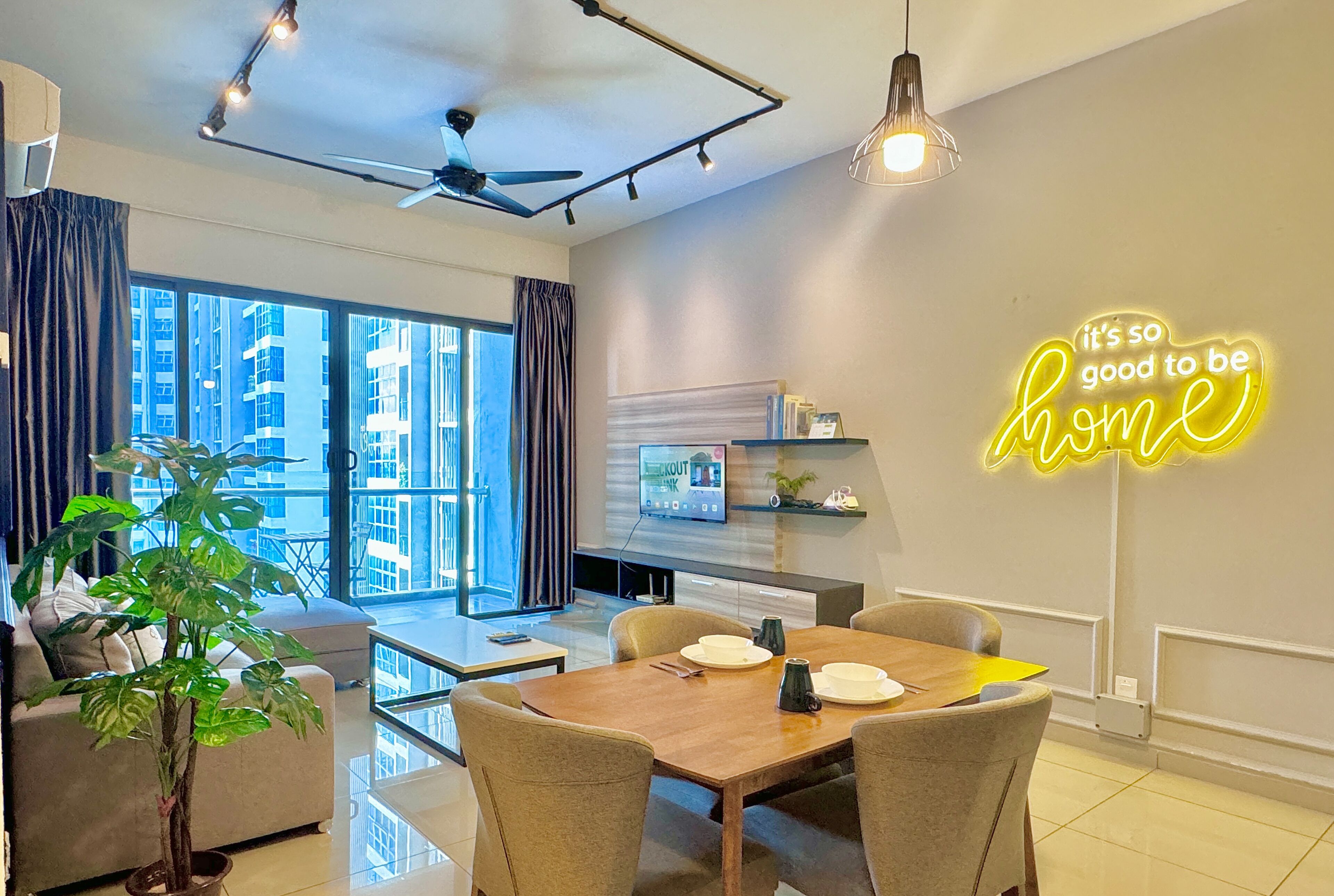 A modern living room with a dining area, large windows, a ceiling fan, and a neon sign on the wall.