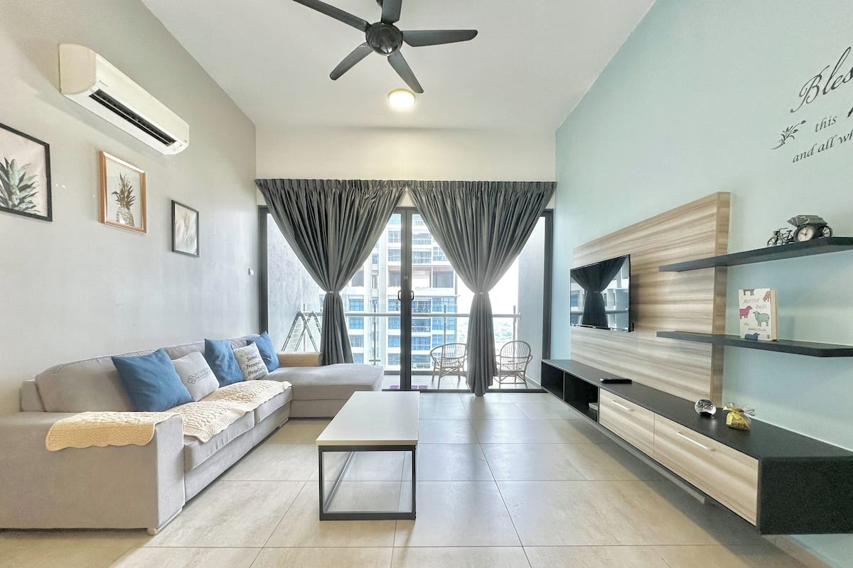 Una sala de estar moderna con sofá, mesa de centro y televisor. Tiene un balcón con cortinas y vista a la ciudad.