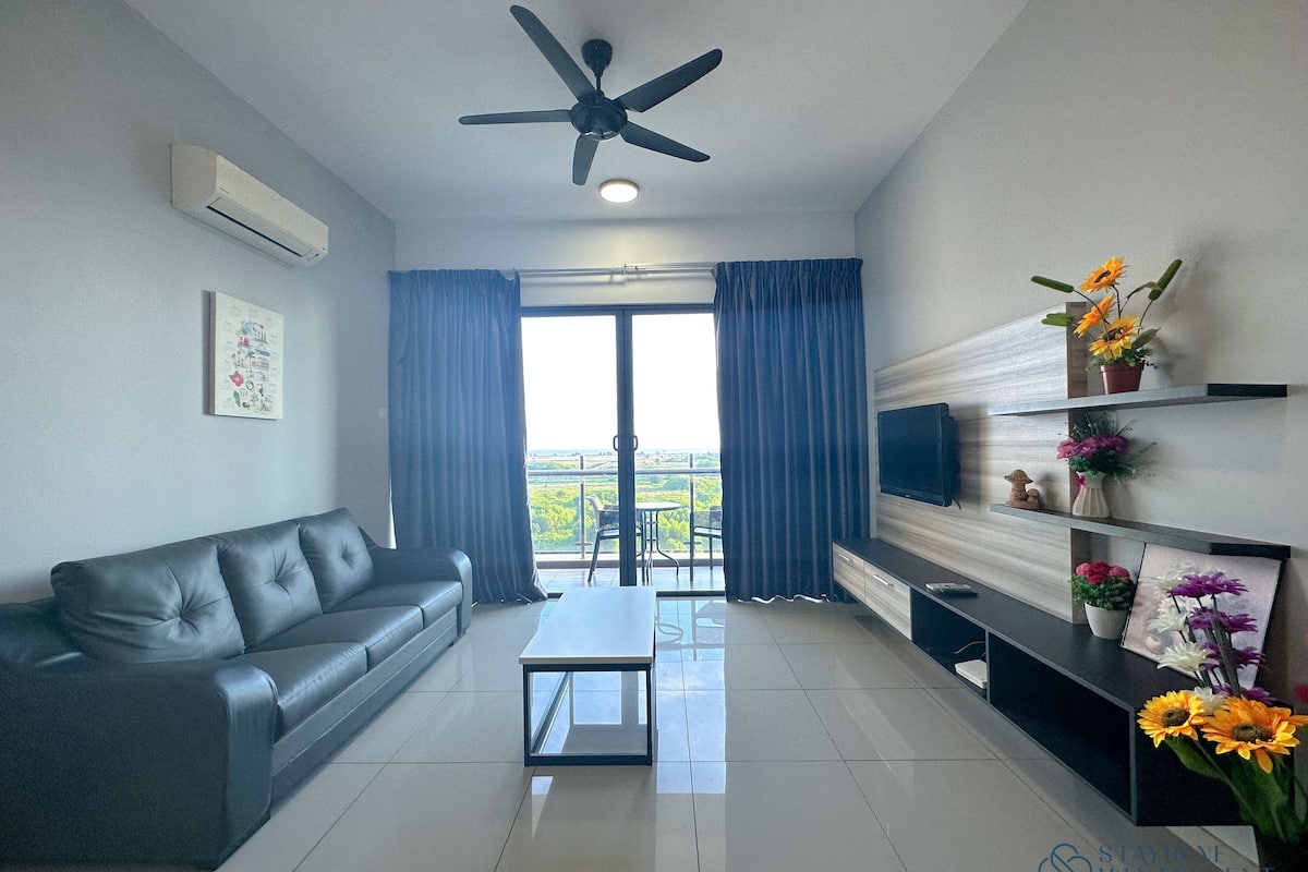 Una sala de estar moderna con un sofá, una mesa de centro y un televisor. Se aprecia un balcón con cortinas y vistas a zonas verdes.
