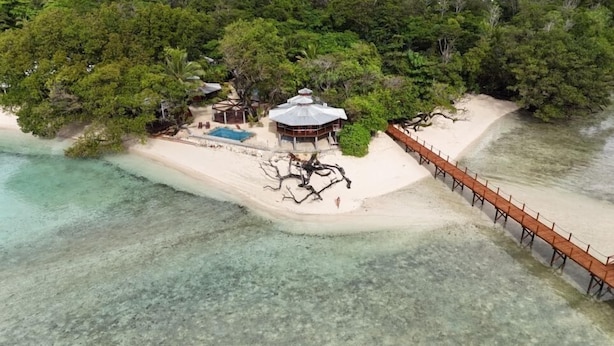 Vista aérea de un complejo turístico frente a la playa, con un muelle de madera, un pabellón y abundante vegetación.