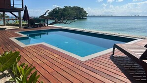 Una piscina rectangular con cubierta de madera, rodeada de una terraza de madera y sillas de descanso, con vista al mar tranquilo y una pequeña isla con árboles.