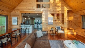 Un vaste chalet de style loft, avec un intérieur en bois, un coin repas doté d’une longue table et de chaises, un salon confortable avec un canapé et une table basse, et une cuisine à l’arrière-plan.