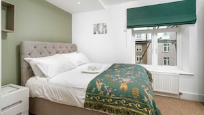 A bedroom with a bed, a nightstand, a window with a view of buildings, and a green curtain. - Stunning 3BD Flat, Parking, Sleeps Six, Near Park (London)