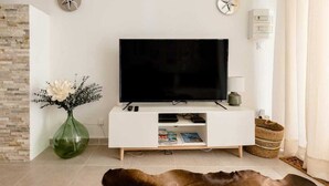 A modern living room with a flat-screen TV on a white entertainment unit, a green vase with flowers, and a stone wall. - Casa Relax Appart Cocooning Dans Village Provençal (Montfort-sur-Argens)