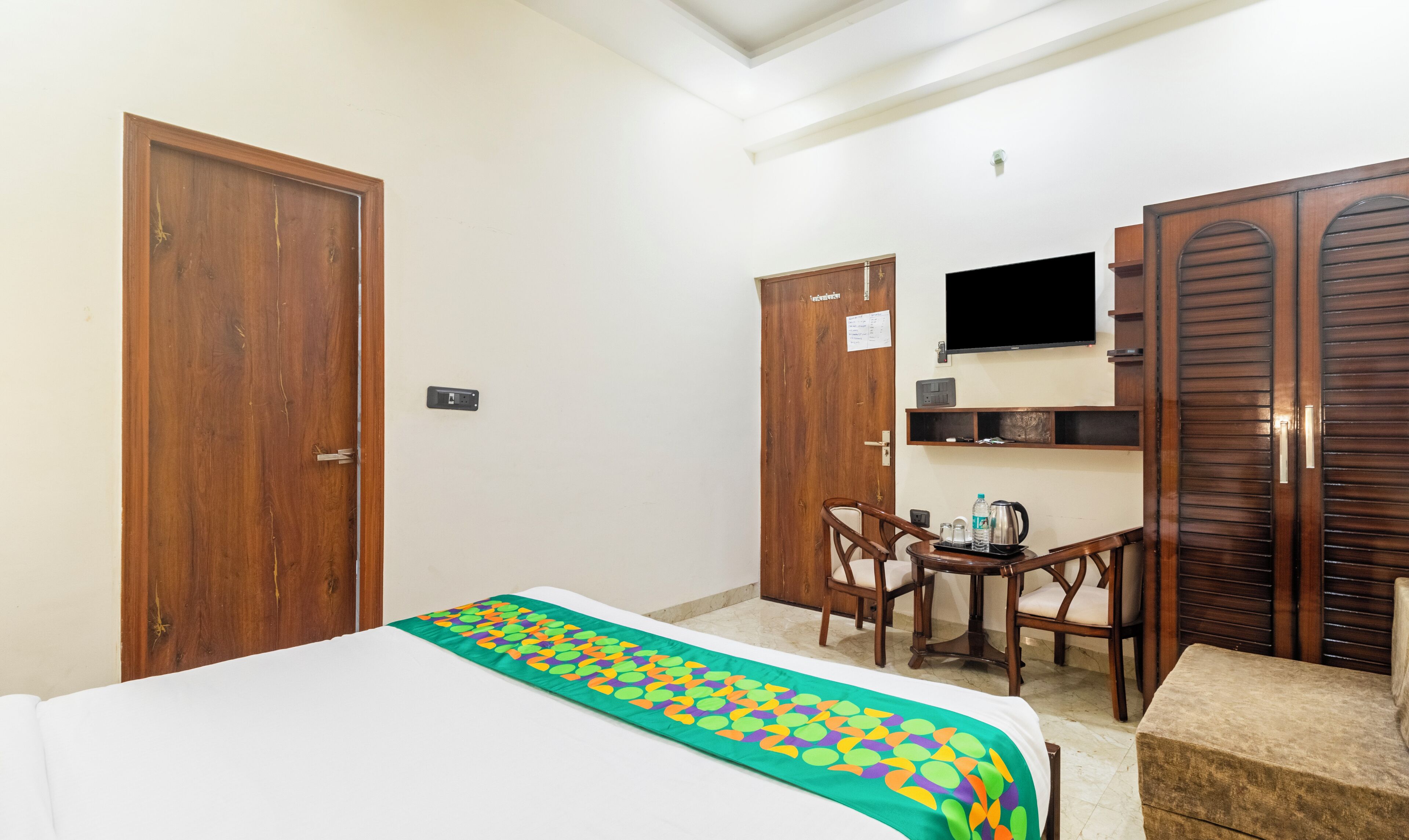 Een hotelkamer met een bed, een bureau met stoel, een aan de muur bevestigde televisie en houten kasten.