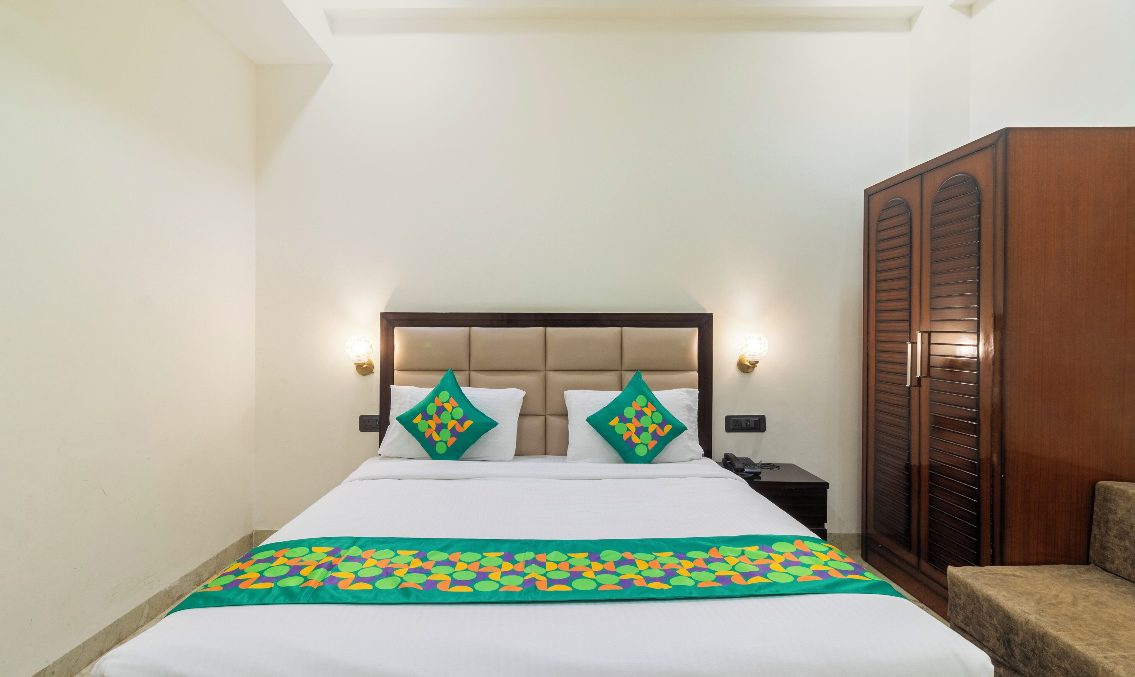 Een hotelkamer met een bed, twee decoratieve kussens, een nachtkastje met een lamp en een kledingkast.