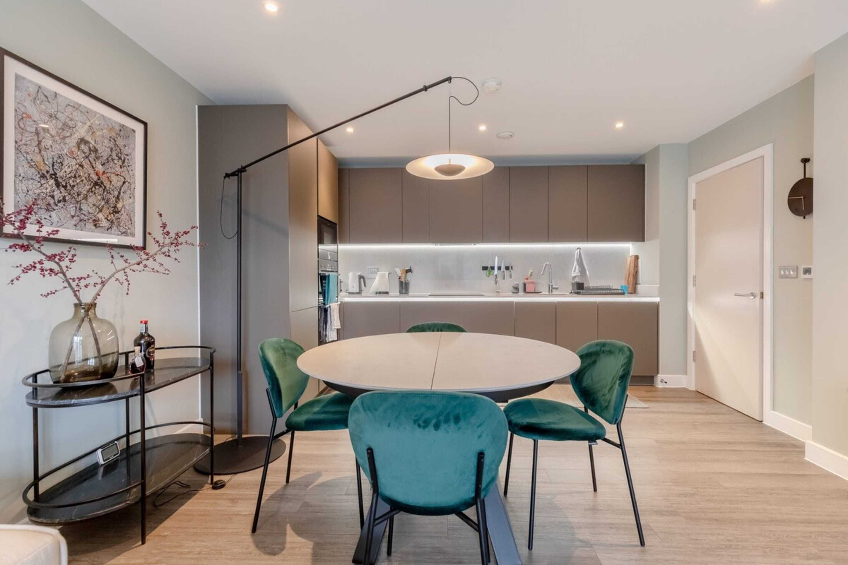 A modern kitchen with a round dining table and teal chairs, a pendant light, and a vase with flowers on a side table.